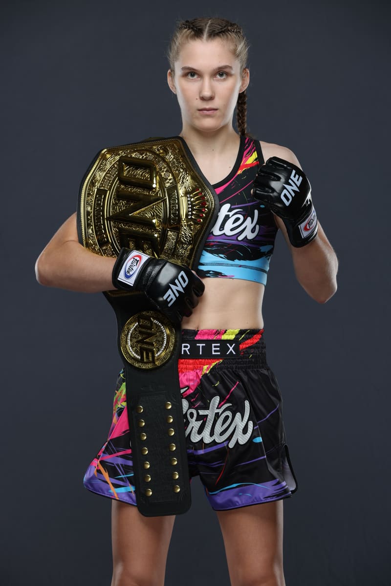 Studio portrait of Smilla Sundell standing with the ONE Championship belt over her shoulder and her gloved fists up, in a black Fairtex fight kit with a colorful paint-splash print