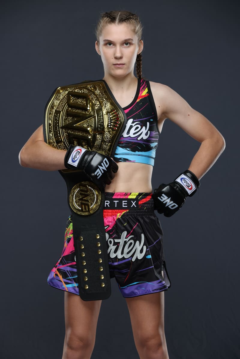 Studio portrait of Smilla Sundell standing with the ONE Championship belt over her shoulder and one hand on her hip, in a black Fairtex fight kit with a colorful paint-splash print