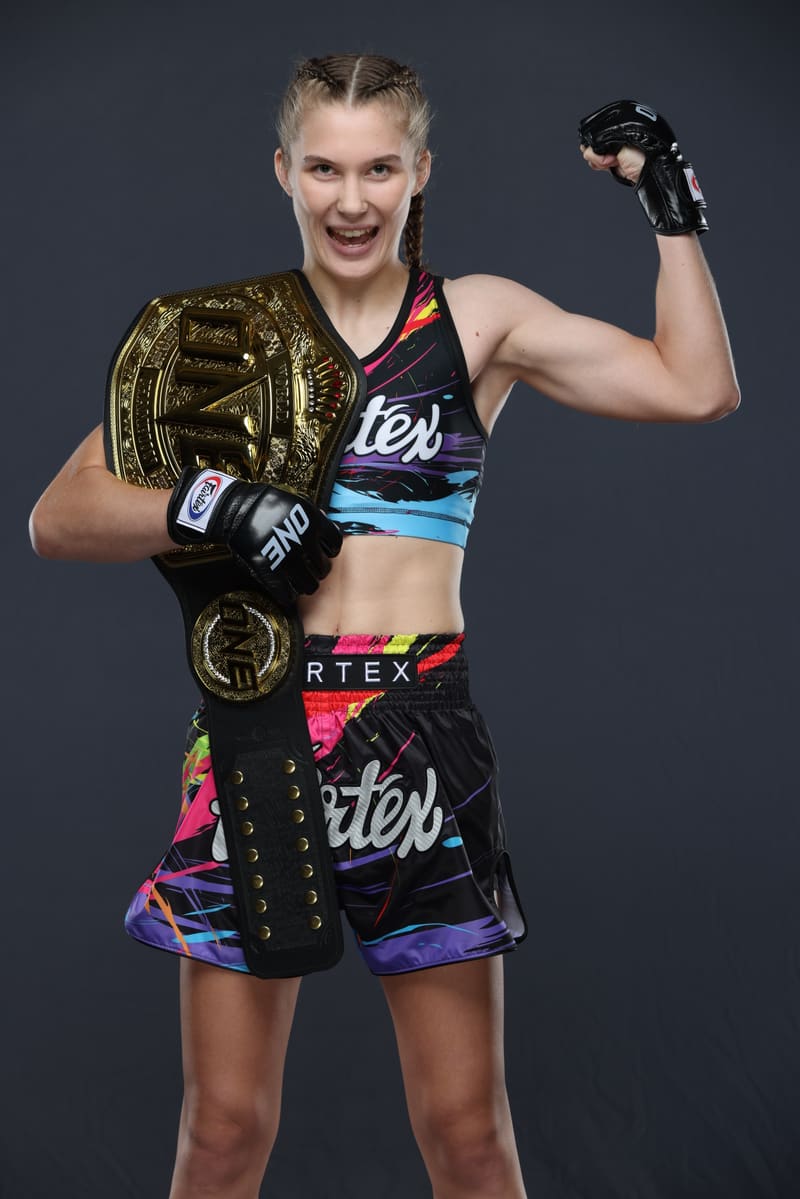 Studio portrait of Smilla Sundell laughing with the ONE Championship belt over her shoulder and one gloved arm raised in a flex, in a black Fairtex fight kit with a colorful paint-splash print