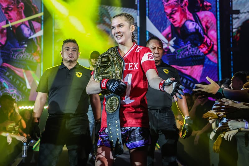 Smilla Sundell walking out to the cage flanked by security staff, carrying the belt over a red walkout jersey