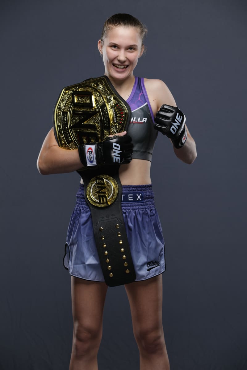 Studio portrait of Smilla Sundell smiling with the ONE Championship belt over her shoulder, in a grey and purple Smilla top and purple Fairtex shorts